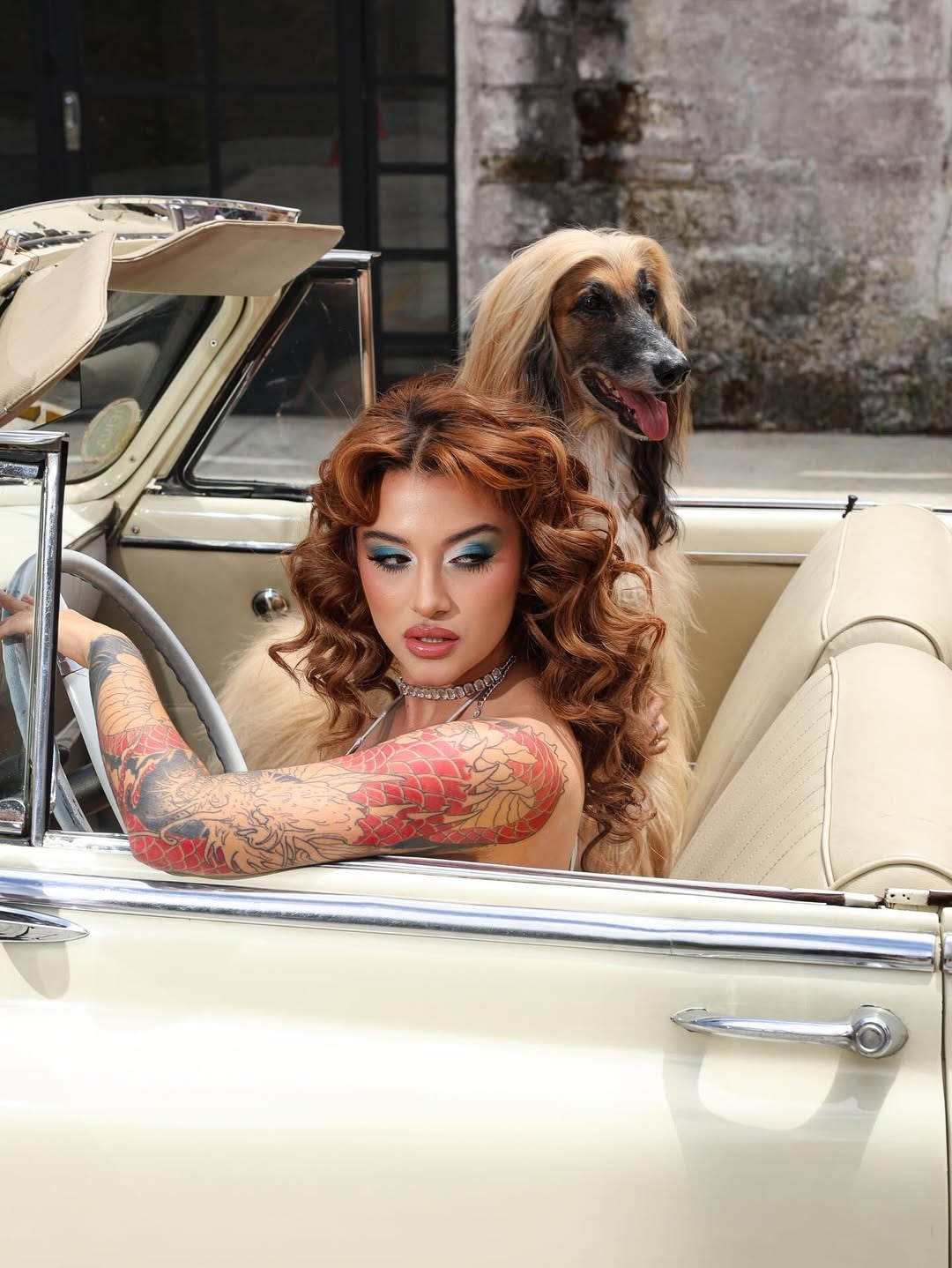 Woman with red curled hair and tattoos in cream convertible with Afghan hound dog