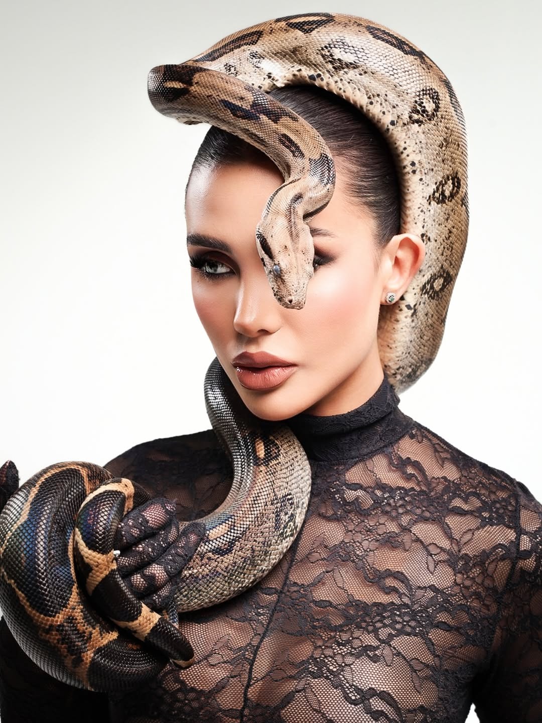 Woman in black lace bodysuit poses with live python draped around shoulders and head against white background
